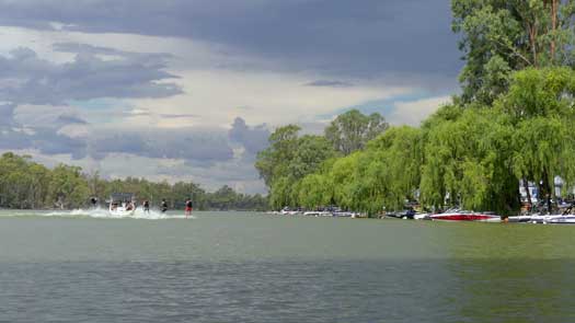 Multiple tow boats line the shore and 5 guys water skiing behind a single boat